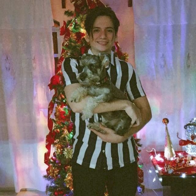 Persona en su sala de estar frente a un árbol de navidad, sosteniendo un perro entre sus brazos, sonriendo a la cámara.